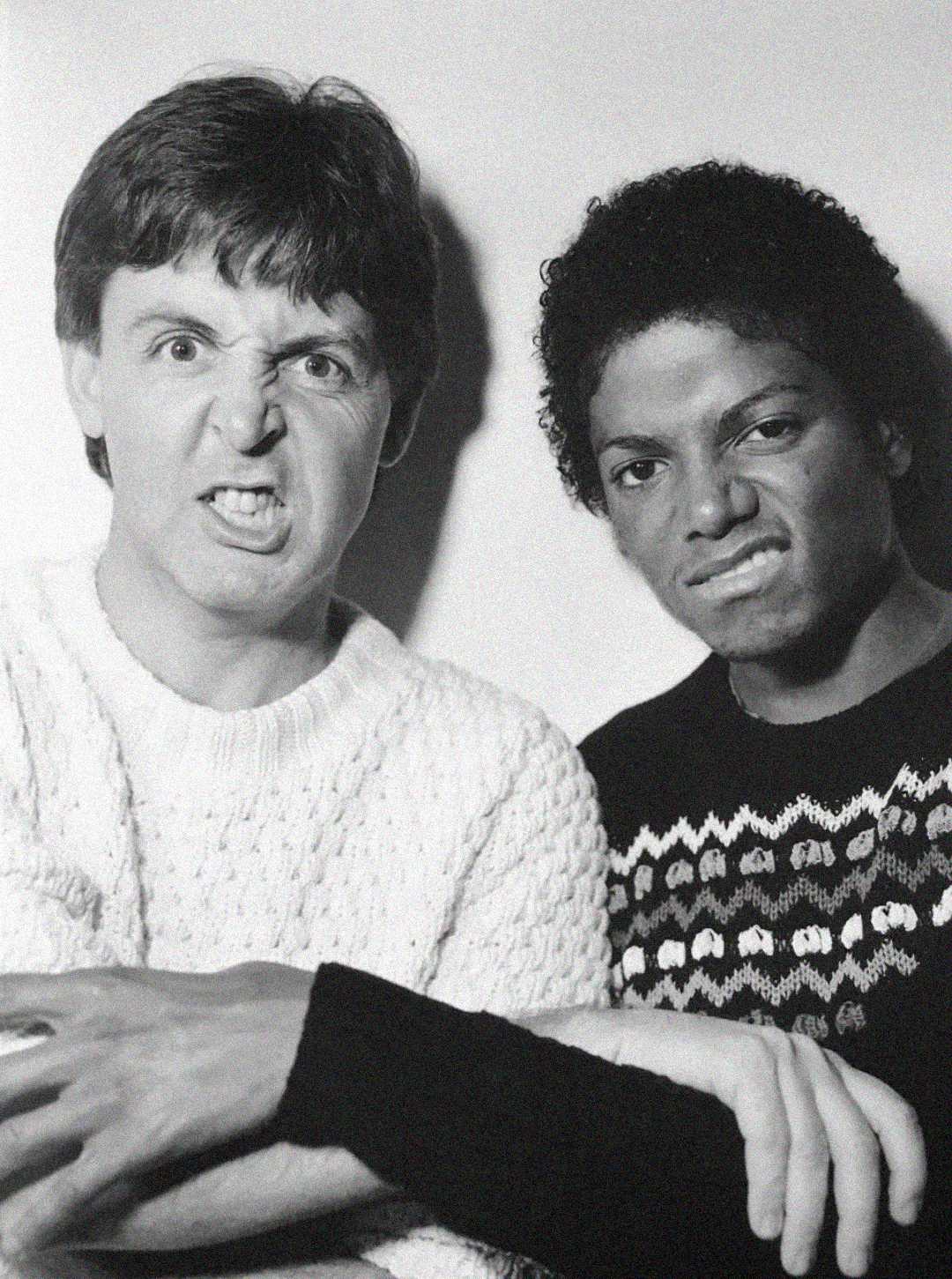 Two men pose together making exaggerated, funny faces; one wears a light knit sweater, the other a dark patterned sweater. The image is black and white.