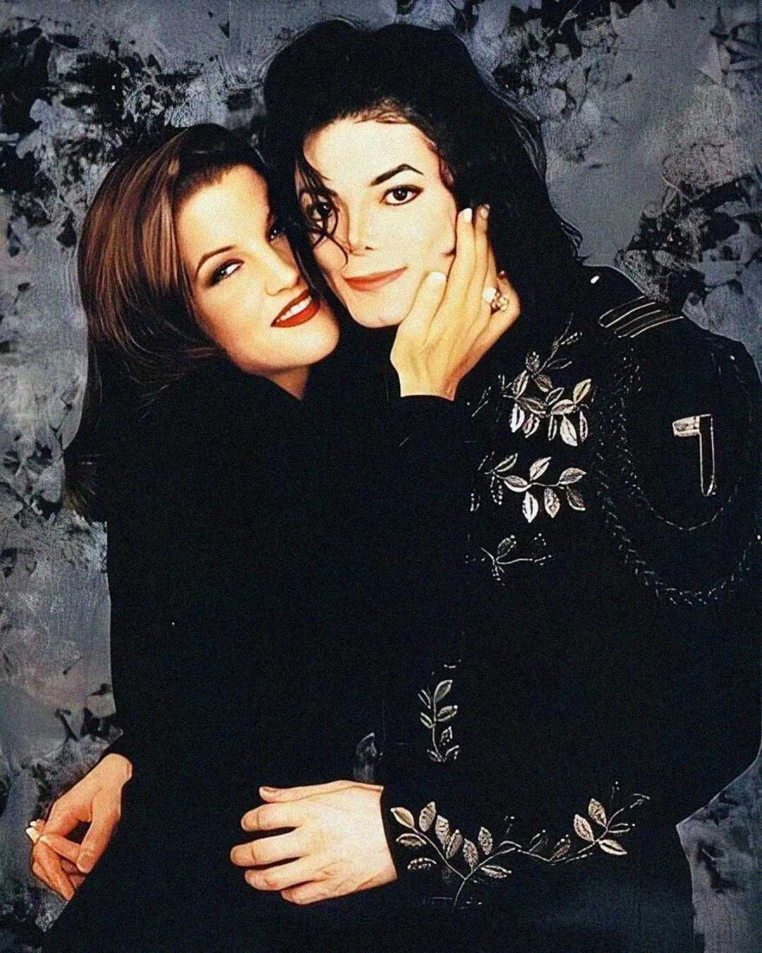 A woman with long brown hair embraces and gently holds the face of a man with long black hair wearing a black embroidered jacket, both smiling in front of a gray, abstract background.