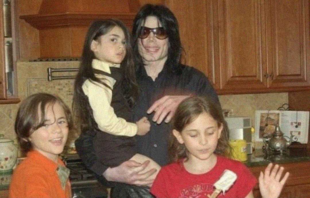 A man in sunglasses stands in a kitchen holding a young girl, with two other children standing beside them. All appear to be posing for a casual photo, and kitchen cabinets and appliances are visible in the background.