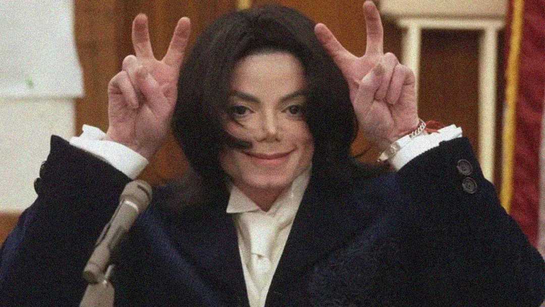 A person with long dark hair and fair skin wearing a dark suit and white shirt is smiling and holding up both hands making peace signs near their head in what appears to be a formal setting.