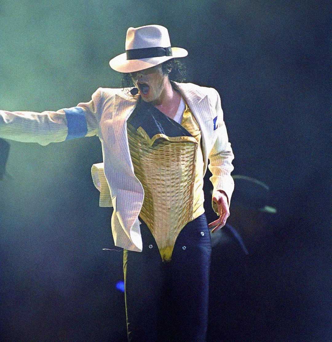 A performer in a white fedora, white pinstripe jacket, gold vest, and black pants dances energetically on stage with one arm outstretched, surrounded by dramatic lighting and haze.