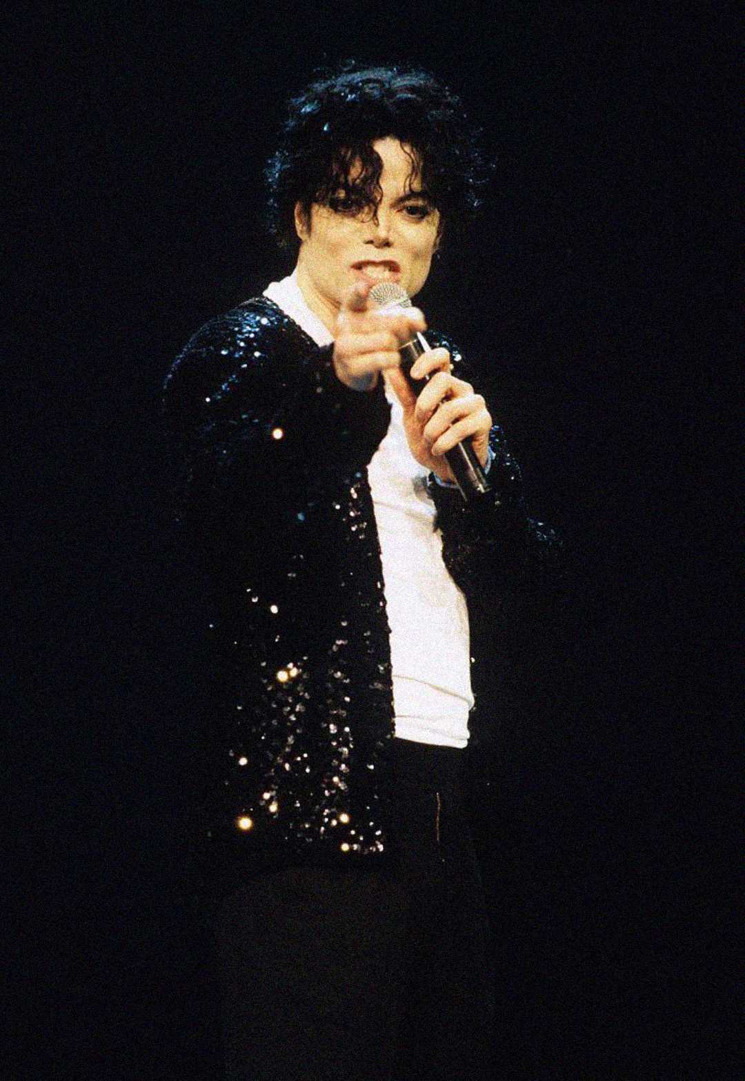 A person with curly dark hair wearing a sequined black jacket and white shirt performs on stage, holding a microphone and pointing towards the audience against a dark background.