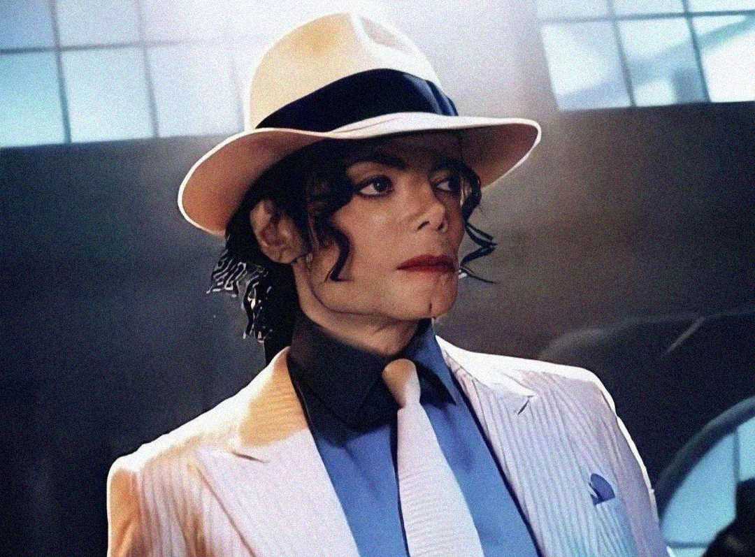 A person wearing a white pinstripe suit, white fedora hat with a black band, light shirt, and white tie stands in a dimly lit room with large windows in the background.