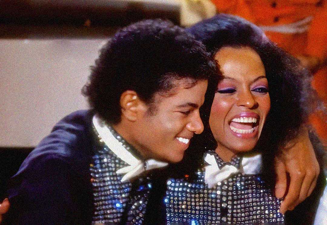 Two people dressed in sparkling outfits smile and embrace, appearing joyful and close. Both wear bow ties and sequined jackets, with one leaning in and the other laughing with mouth open.