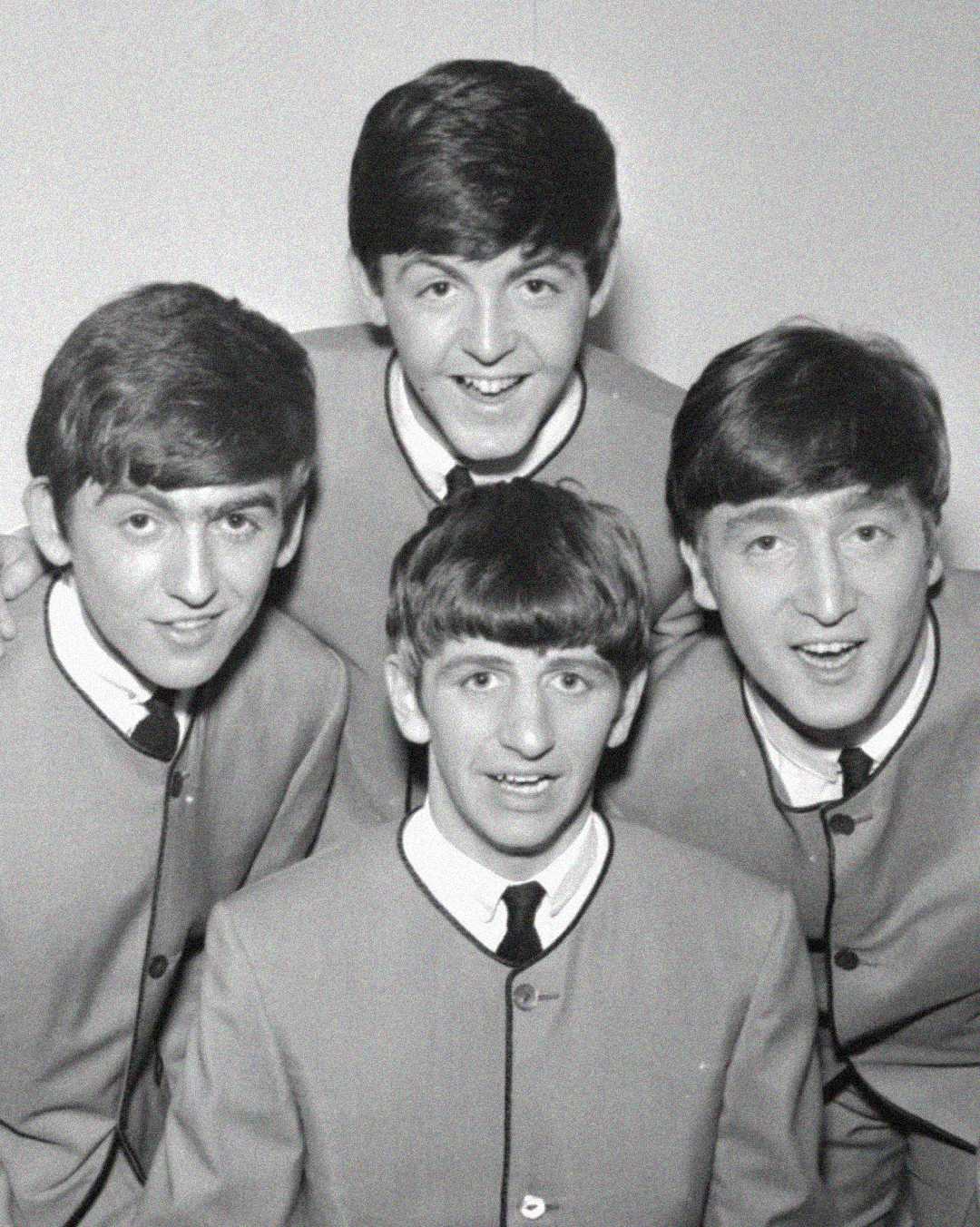 Four young men wearing matching suits pose closely together and smile at the camera in a black-and-white photo.
