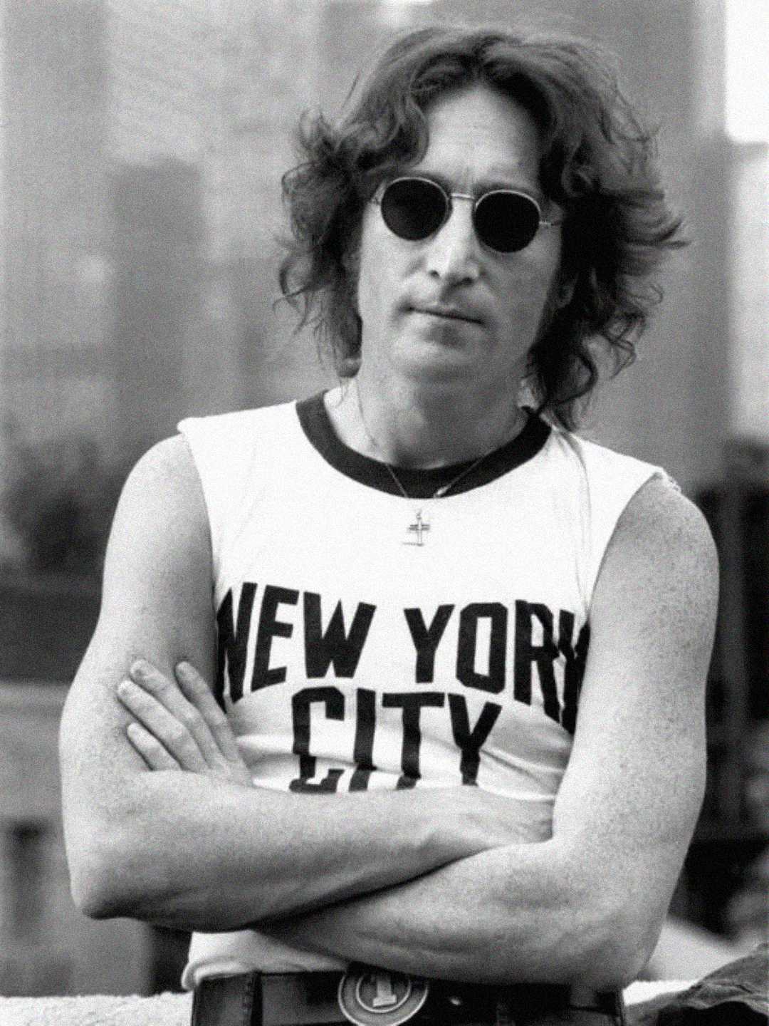 A person with medium-length wavy hair wearing round sunglasses and a sleeveless shirt that says "New York City" stands with arms crossed. The background appears to be an urban setting.