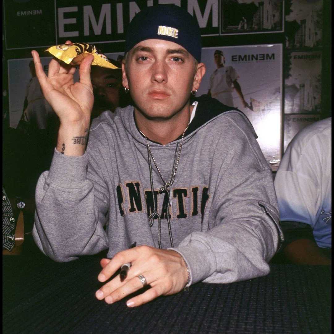 A man wearing a blue cap backward and a gray hoodie sits at a table, holding a yellow object in one hand. He has a serious expression and several chains around his neck. Posters are visible in the background.