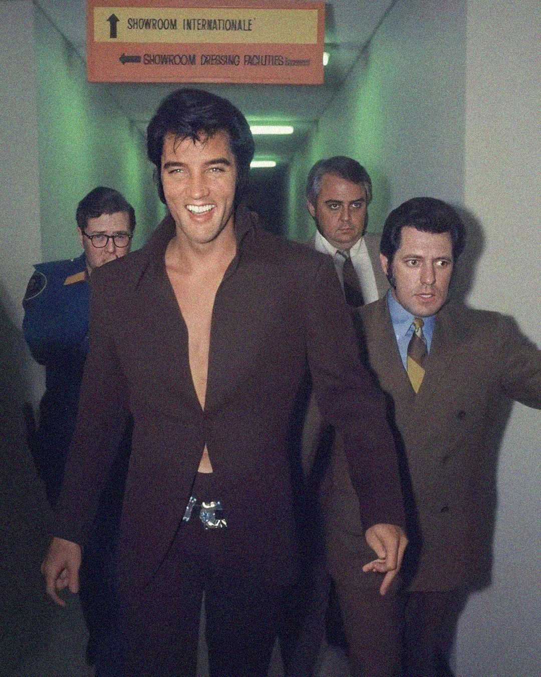A man with dark hair and sideburns, wearing a brown suit with an open collar and a large belt buckle, smiles as he walks down a hallway, accompanied by several serious-looking men in suits.
