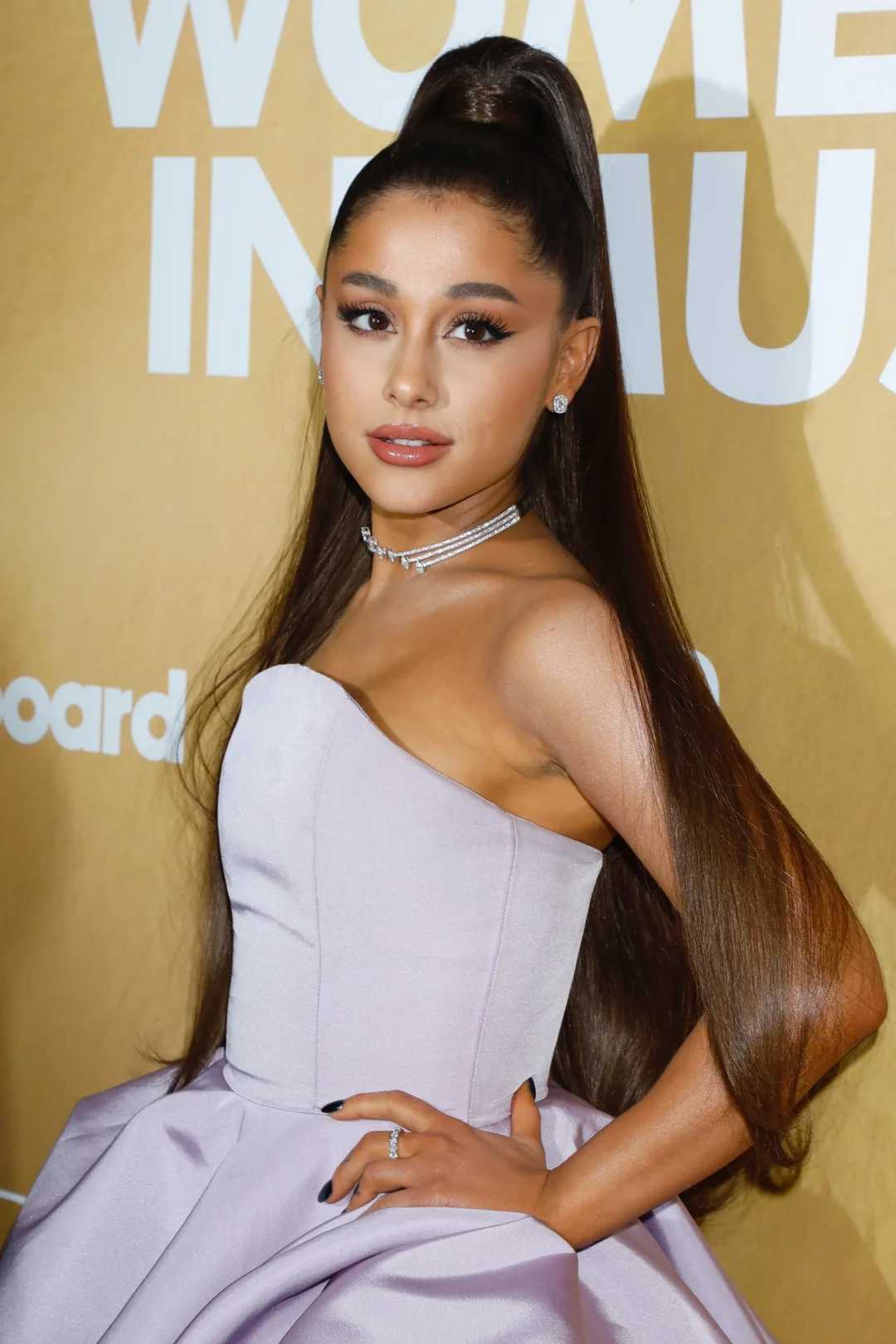 A woman with long, straight brown hair in a high ponytail wears a strapless, light lavender gown and a layered necklace, posing on a red carpet in front of a gold backdrop with white text.