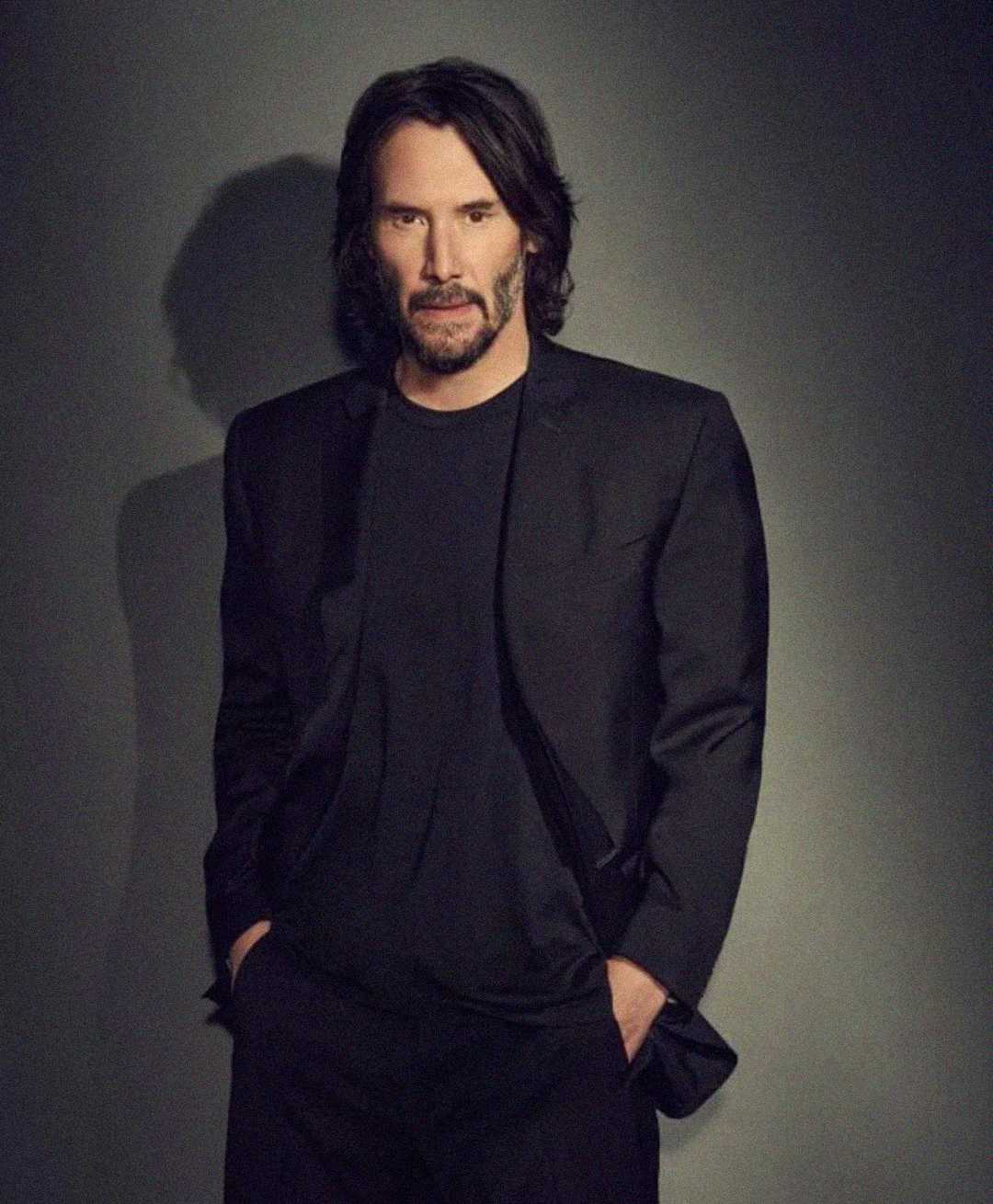 A man with shoulder-length dark hair and a beard stands against a plain, dark background wearing a black suit and black shirt, with his hands in his pockets and a neutral expression.