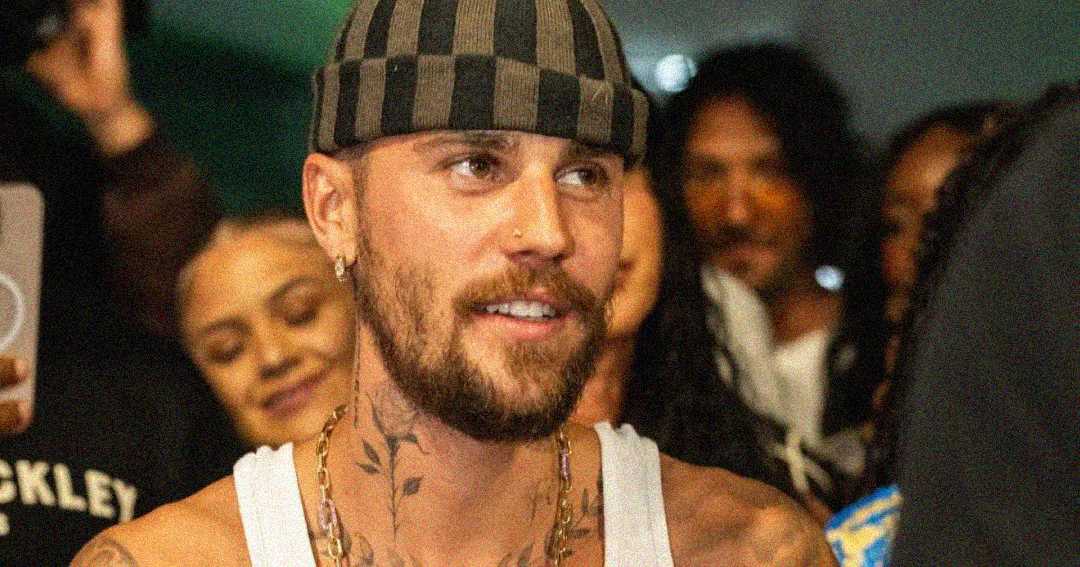 A bearded man wearing a striped beanie and white tank top smiles while surrounded by a crowd of people in the background.
