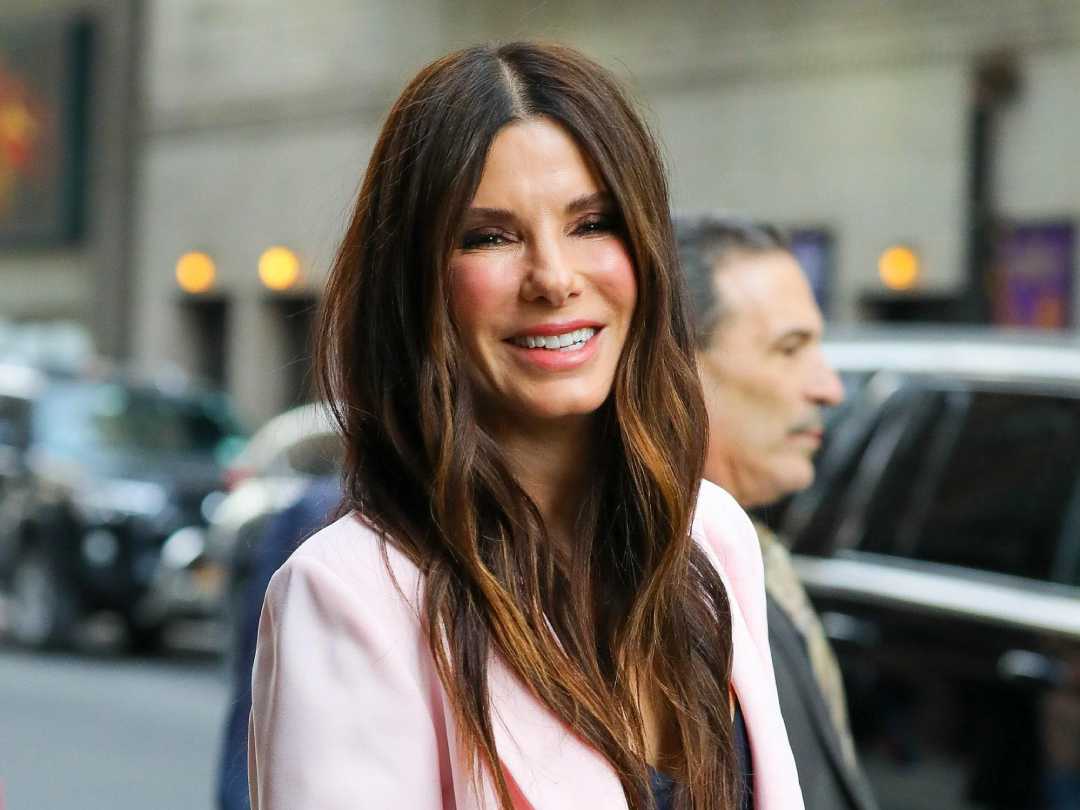 A woman with long brown hair and a light pink blazer smiles while standing outdoors on a city street, with blurred people and cars in the background.