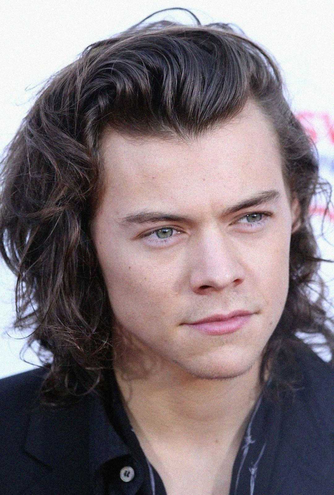 A young person with shoulder-length wavy brown hair and blue eyes looks slightly to the side. They have light skin and a calm expression, wearing a dark jacket over a dark shirt. The background is bright and out of focus.