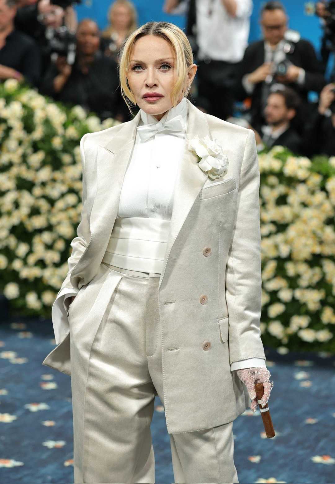 A woman wearing a cream-colored tuxedo with a white shirt, bowtie, flower lapel pin, and gloves stands confidently on a floral carpet, with photographers and a wall of white flowers in the background.