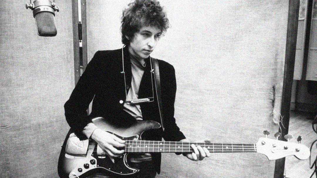 A black and white photo of a young man with curly hair playing an electric bass guitar in a recording studio, wearing a jacket and a harmonica holder around his neck. A large microphone hangs nearby.