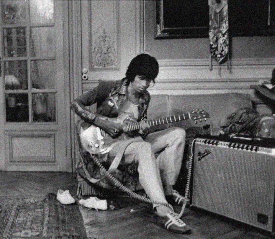 Black and white photo of a person sitting on a couch playing an electric guitar, with an amplifier nearby. They are wearing a jacket, light pants, and sneakers, with a shoe off and cables on the floor in a vintage room.