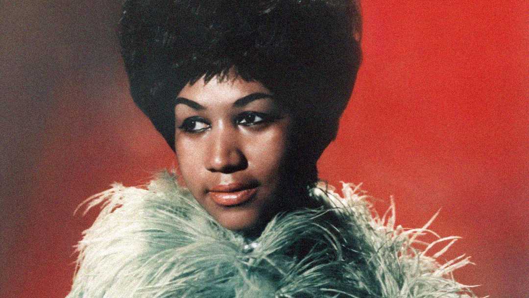 A woman with styled dark hair wears a light green feathered garment, gazing slightly to the side against a gradient red background.