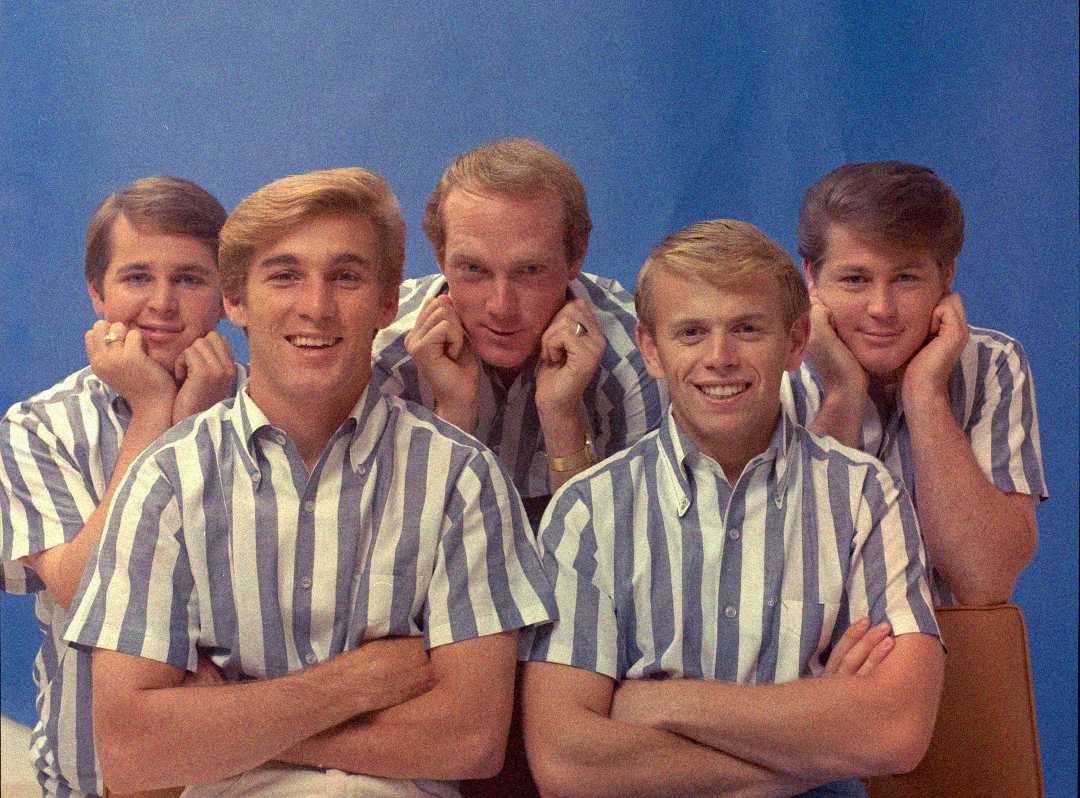 Five young men wearing matching blue-and-white striped shirts pose together in front of a plain blue background, smiling and resting their chins on their hands.