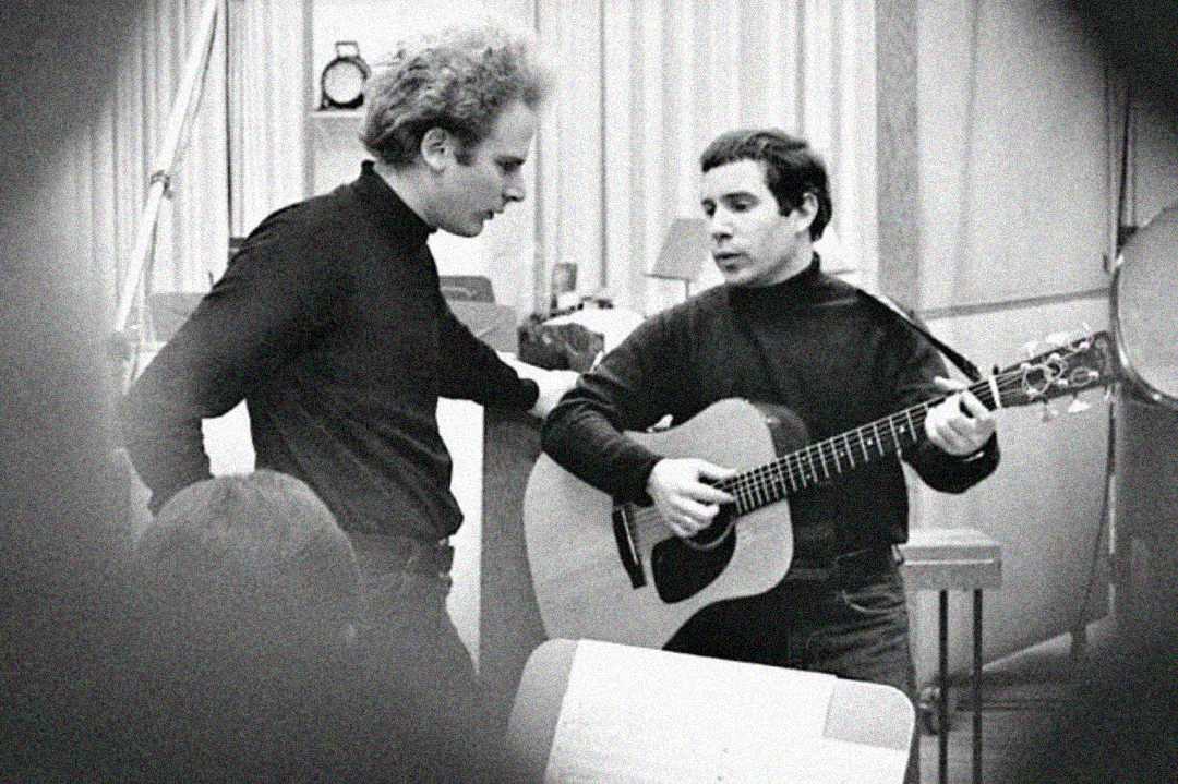 Two men in black turtlenecks stand in a recording studio; one is playing an acoustic guitar while the other listens intently. The scene is black and white, with studio equipment visible in the background.