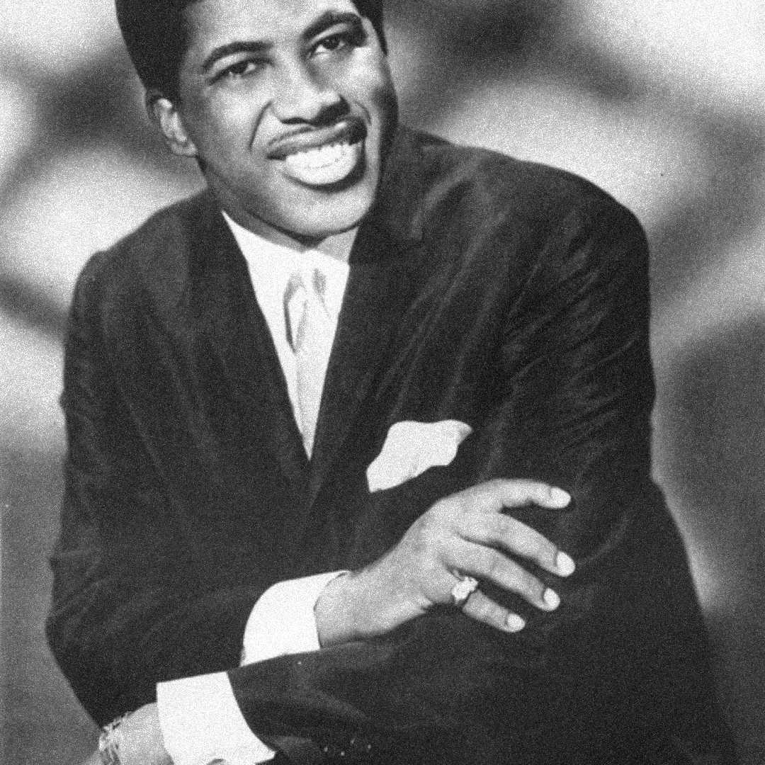 A smiling man in a dark suit, white shirt, and tie poses with his arms crossed. He wears a ring and watch, and a handkerchief is visible in his breast pocket. The background is softly blurred. The image is in black and white.