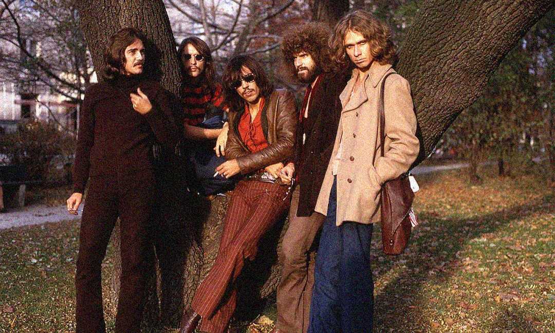 Five men in 1970s-style clothing stand and lean against a large tree in a park, surrounded by autumn leaves, posing for a relaxed group photo.