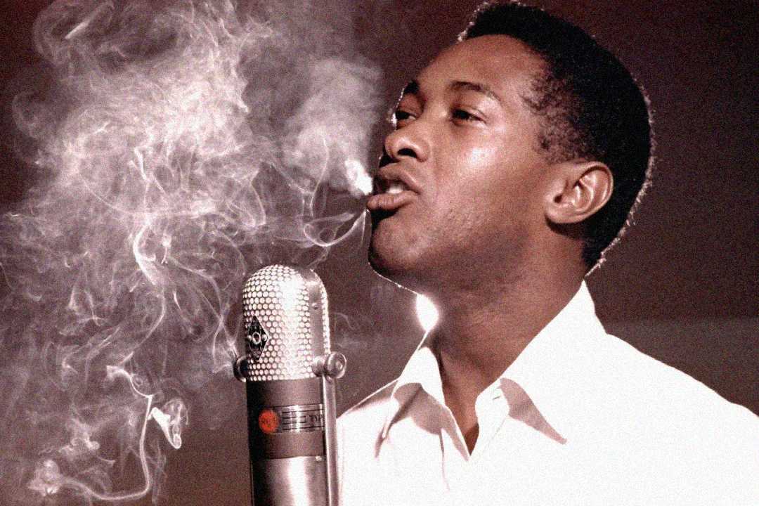 A man in a white shirt sings passionately into a vintage microphone as smoke swirls around him, creating a dramatic and atmospheric scene.
