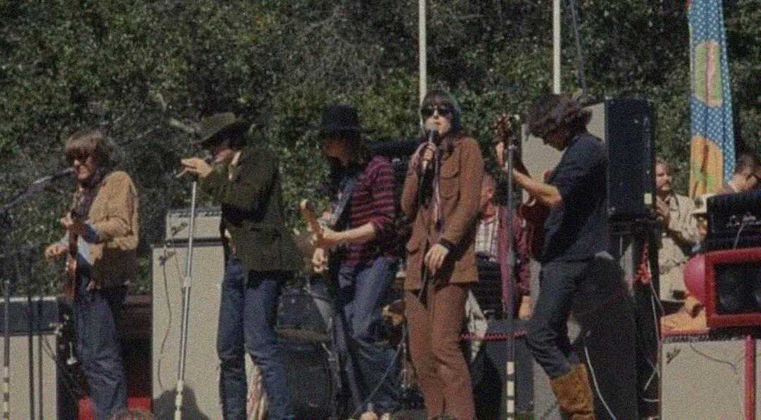 A vintage photo of a band performing on an outdoor stage. Five musicians stand in a row with instruments under daylight, trees in the background, and an audience watching from the side.