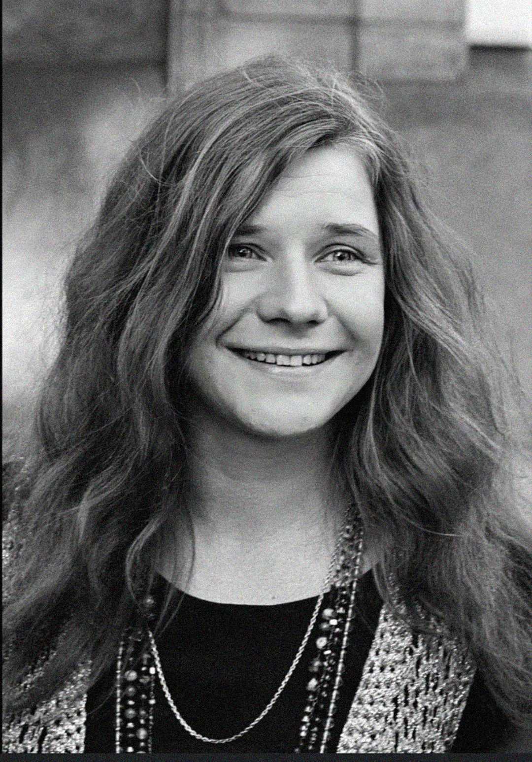 A young woman with long, wavy hair smiles at the camera. She is wearing a dark top, layered necklaces, and a patterned vest. The background is out of focus.