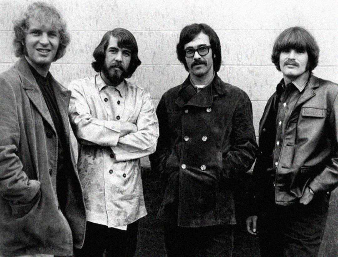 Four men standing side by side in front of a plain wall, wearing 1960s-style jackets and shirts, with long hair and facial hair, looking at the camera, one smiling. Image is in black and white.