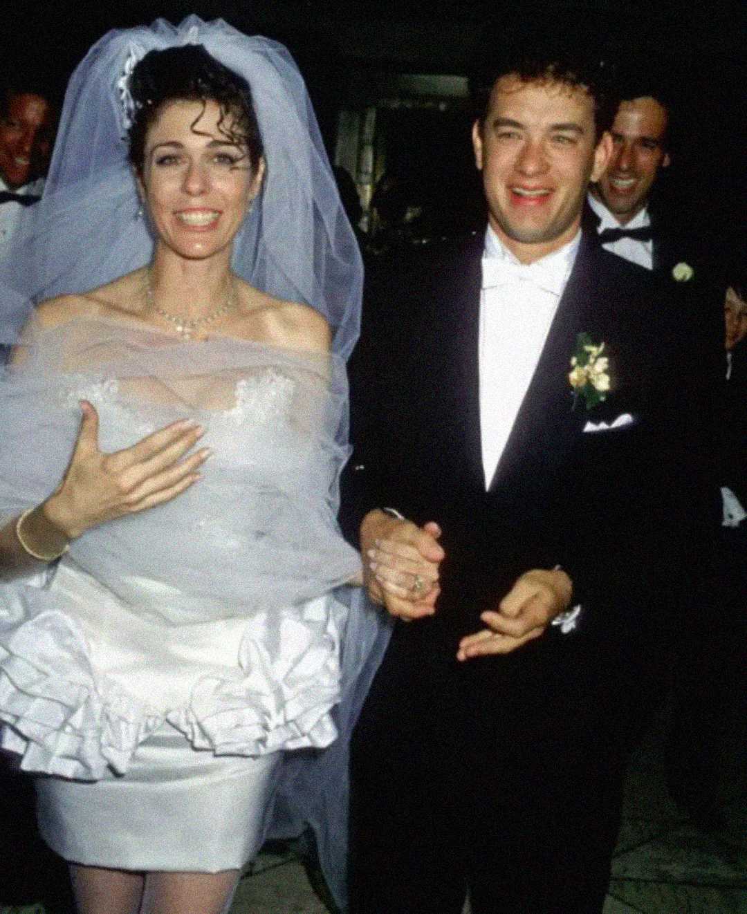 A bride in a white dress and veil and a groom in a black tuxedo smile and hold hands while walking, surrounded by people in formal attire.