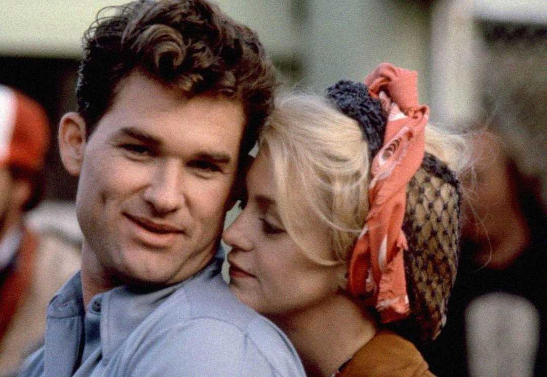 A man with dark hair smiles while a blonde woman wearing a patterned scarf and headband leans affectionately on his shoulder, both appearing happy and relaxed outdoors.