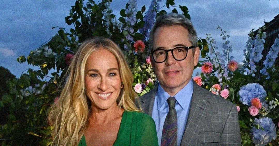 A smiling woman in a green dress poses next to a man in glasses and a gray plaid suit, with a lush background of flowers and greenery under a twilight sky.