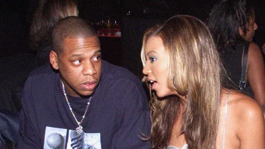 A man wearing a dark shirt and chain necklace sits next to a woman with long, light brown hair, as she leans in to talk to him in a dimly lit, crowded setting.