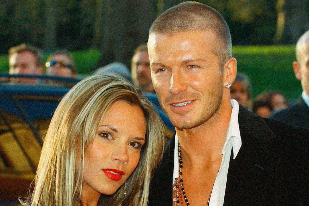 A woman with long blonde hair and bright red lipstick stands next to a man with short hair and facial stubble. Both are dressed formally and smiling, with blurred people and greenery in the background.