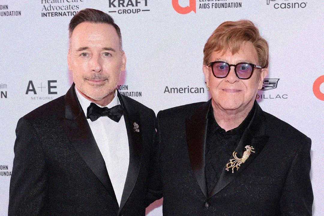 Two men in black tuxedos pose together on a red carpet. One wears glasses and both have pins on their lapels. Step-and-repeat banner with logos from sponsors is visible in the background.