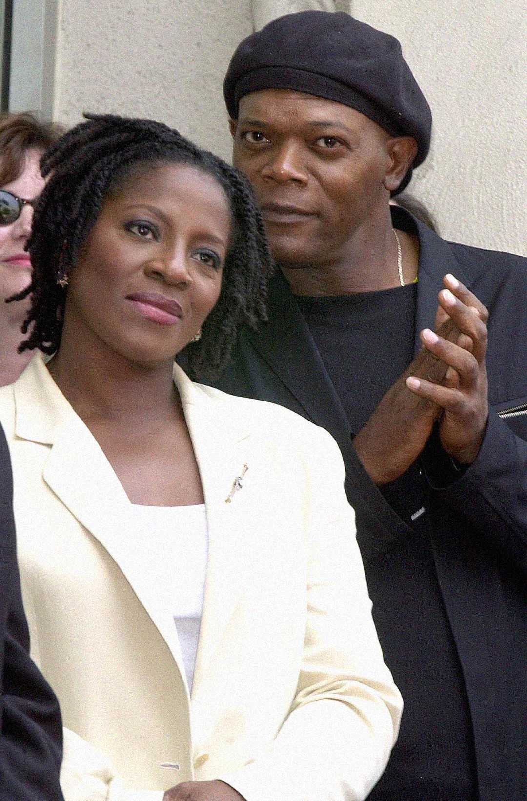 A woman in a white blazer stands in front, smiling softly, while a man in a black outfit and beret stands behind her, hands clasped, looking forward. Both appear calm and attentive.
