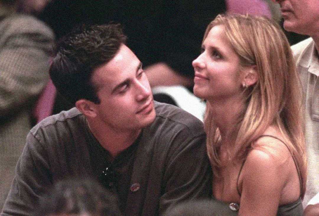 A man and woman sit closely together in a crowd. The man looks lovingly at the smiling woman, who gazes slightly upward. Both appear relaxed and happy, with other people blurred in the background.