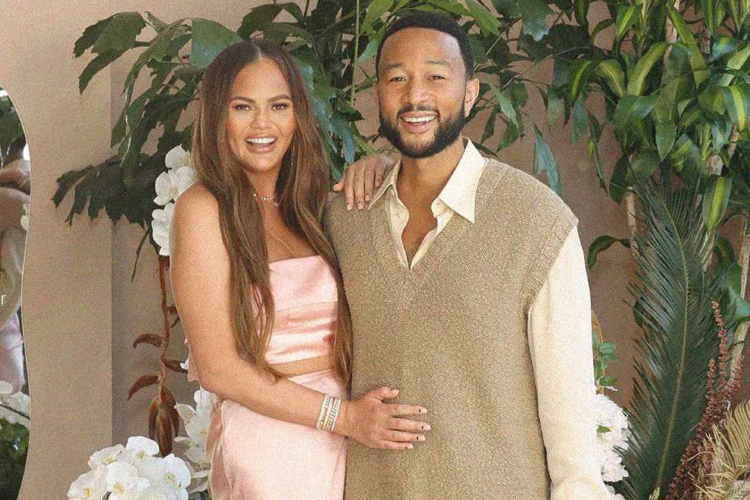 A smiling couple poses together in front of lush green plants and white flowers. The woman wears a pale pink dress, and the man wears a beige shirt with a textured vest. They stand close, with her hand resting on his chest.