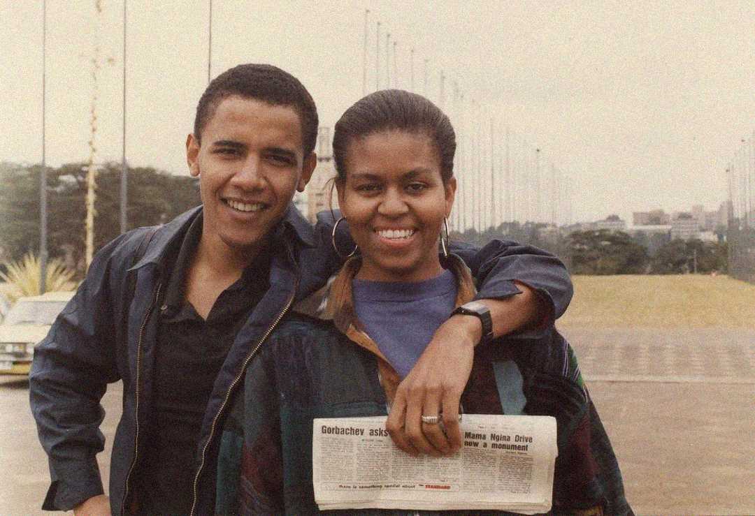A young couple smiles at the camera outdoors; the woman holds a newspaper, and the man stands with his arm around her shoulders. A tree-lined path and flag poles are visible in the background.