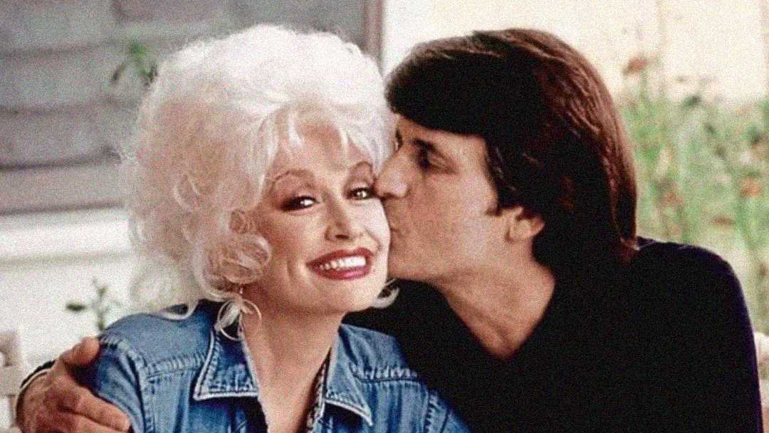 A smiling woman with voluminous white hair and a denim jacket is hugged and kissed on the cheek by a man with dark hair in a cozy, light-filled setting.