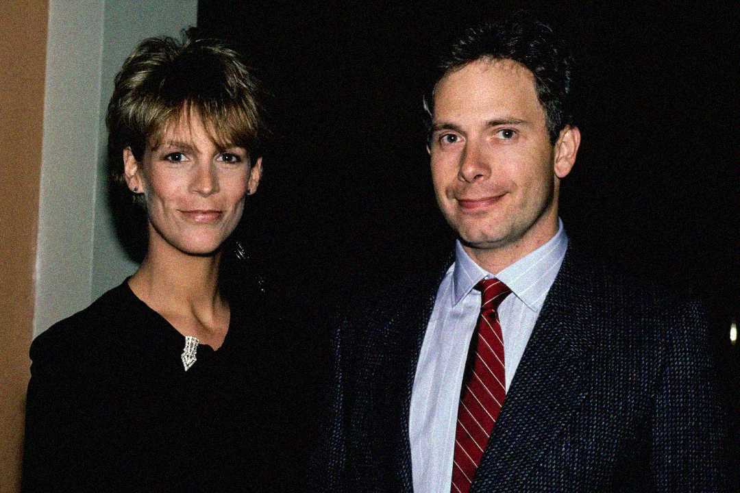 A woman with short hair and a man in a suit and red striped tie stand together indoors, both smiling slightly at the camera against a dark background.