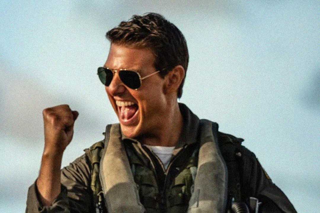 A man in a flight suit and aviator sunglasses pumps his fist and cheers enthusiastically, with his mouth open in excitement against a blurred outdoor background.