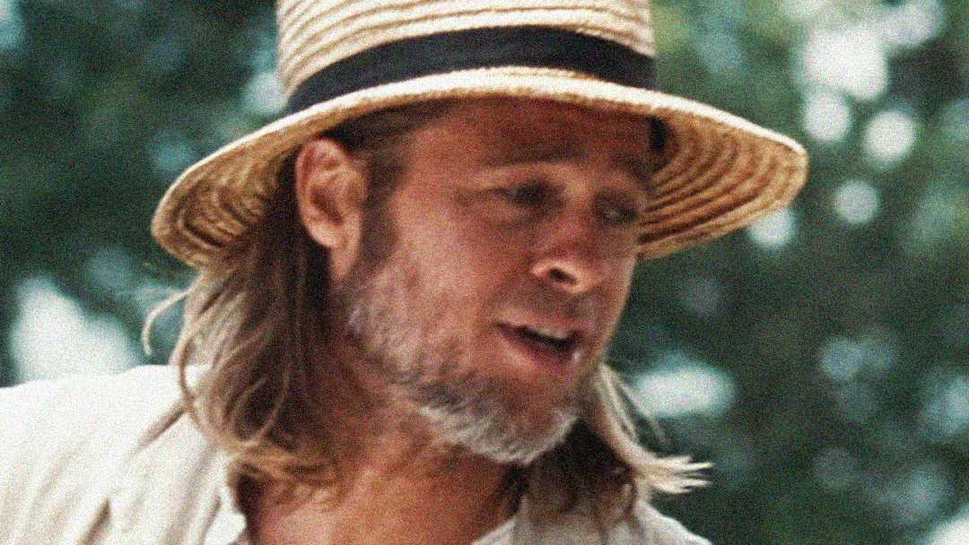 A man with long hair and a beard wears a straw hat with a black band. He is outdoors with blurred greenery in the background and appears to be speaking or singing.