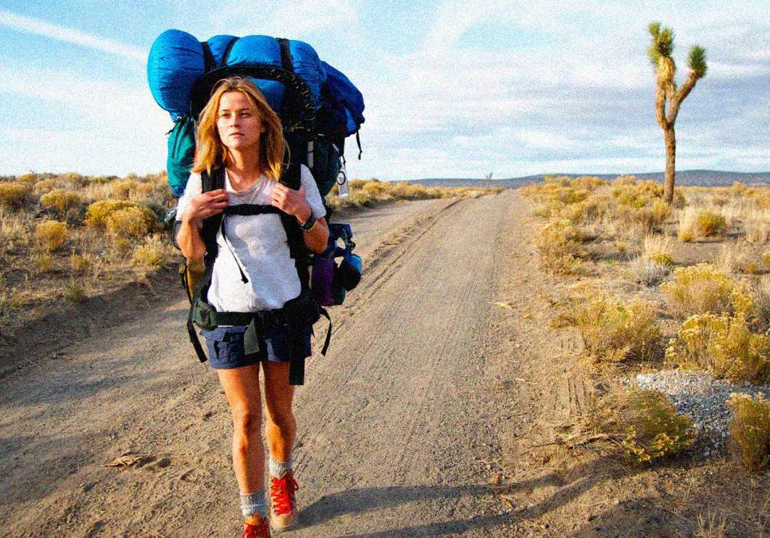 A woman with blonde hair, wearing a white shirt and shorts, hikes on a dirt road in a desert landscape, carrying a large blue and green backpack. Dried bushes and a lone tree are visible under a partly cloudy sky.
