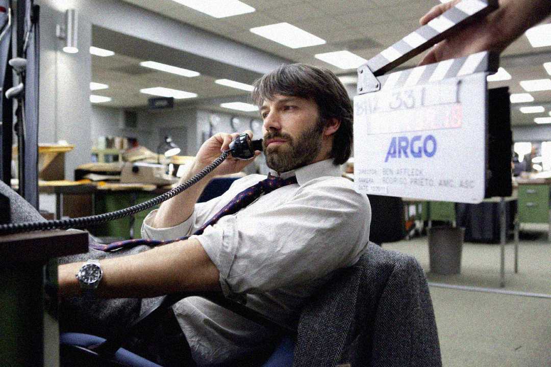A man with dark hair and a beard sits at a desk in an office, talking on a corded phone. A movie clapperboard labeled "ARGO" is held in front of him, suggesting a film set.