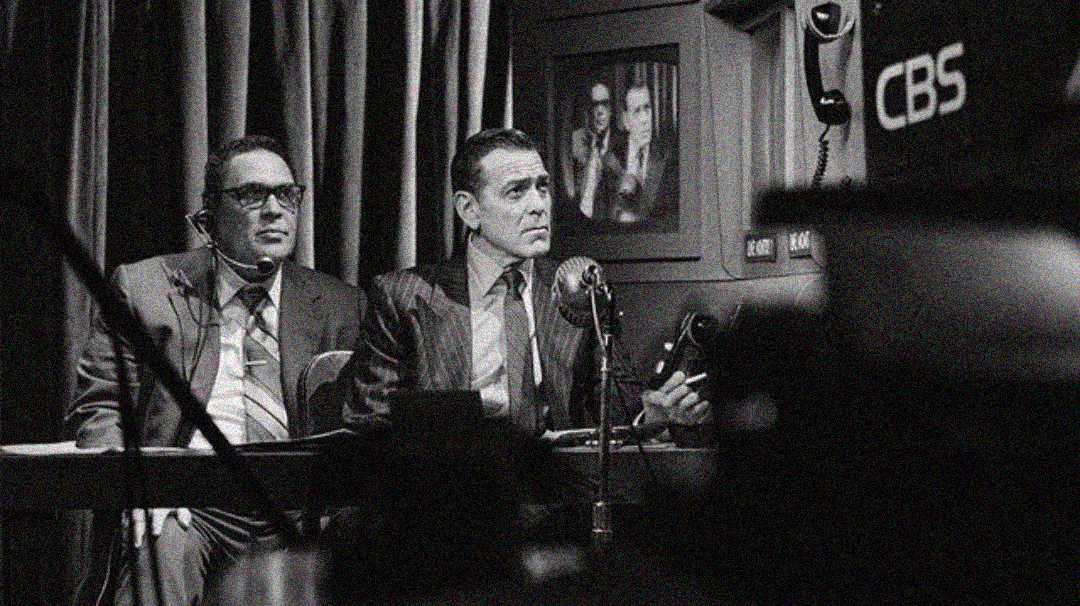 Two men in suits are seated at a desk with microphones and headphones, appearing to commentate or broadcast. Behind them, there’s a framed photo on the wall. A CBS camera is visible in the right foreground. The image is black and white.