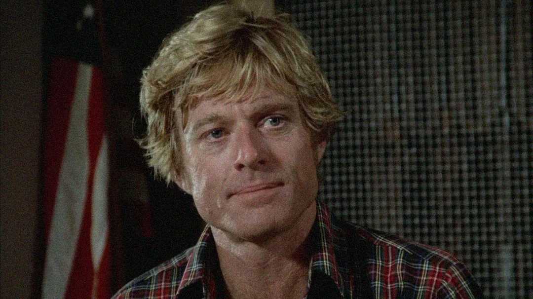 A man with wavy blond hair wearing a red and black plaid shirt sits indoors, looking slightly to the side. An American flag is partially visible in the background.
