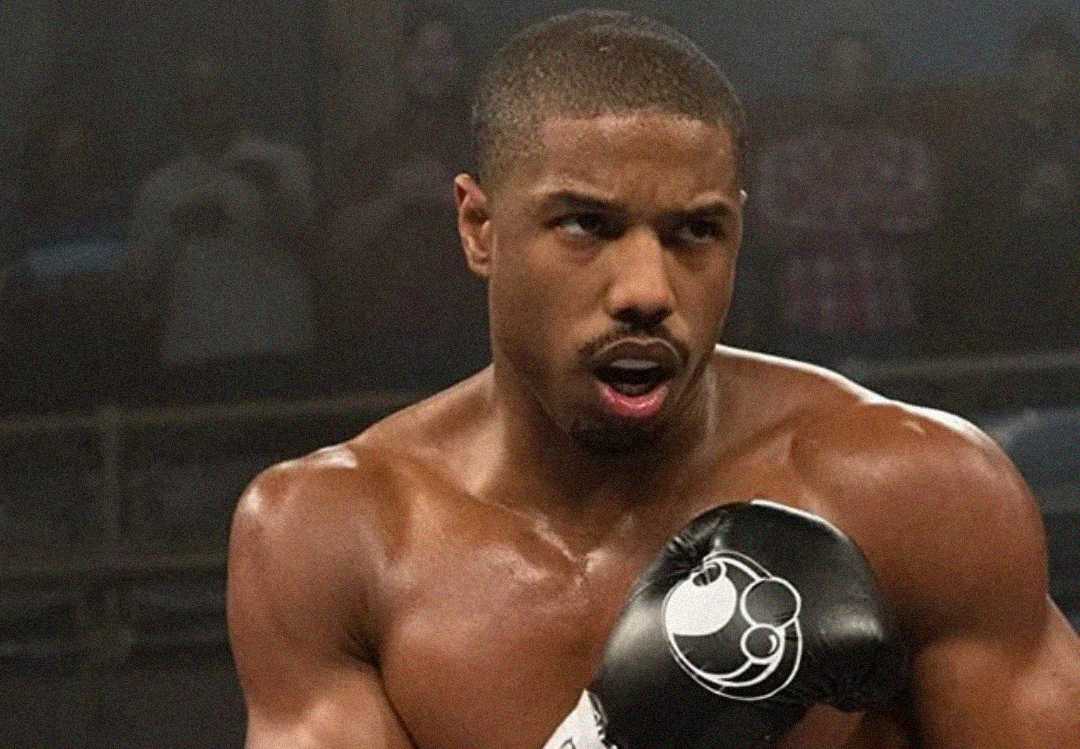 A muscular man wearing boxing gloves stands in a boxing ring, looking focused and determined. His bare torso is glistening with sweat as he prepares to throw a punch. The background is dimly lit, with blurred spectators.