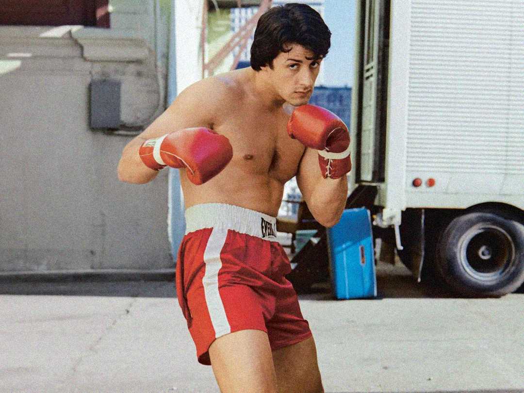 A man wearing red boxing gloves, red shorts with white stripes, and no shirt stands in a boxing stance outdoors near a white vehicle.