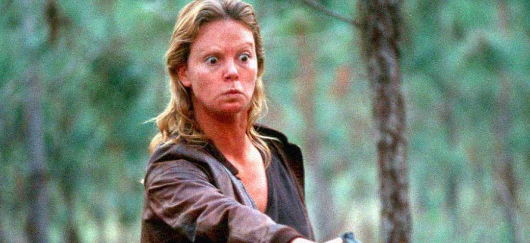 A woman with wide, intense eyes and a serious expression stands in a forest, wearing a brown jacket and pointing a gun forward. The background is blurred with green trees.